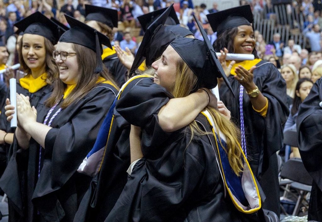 Two graduates hug, amid a large group of graduates.
