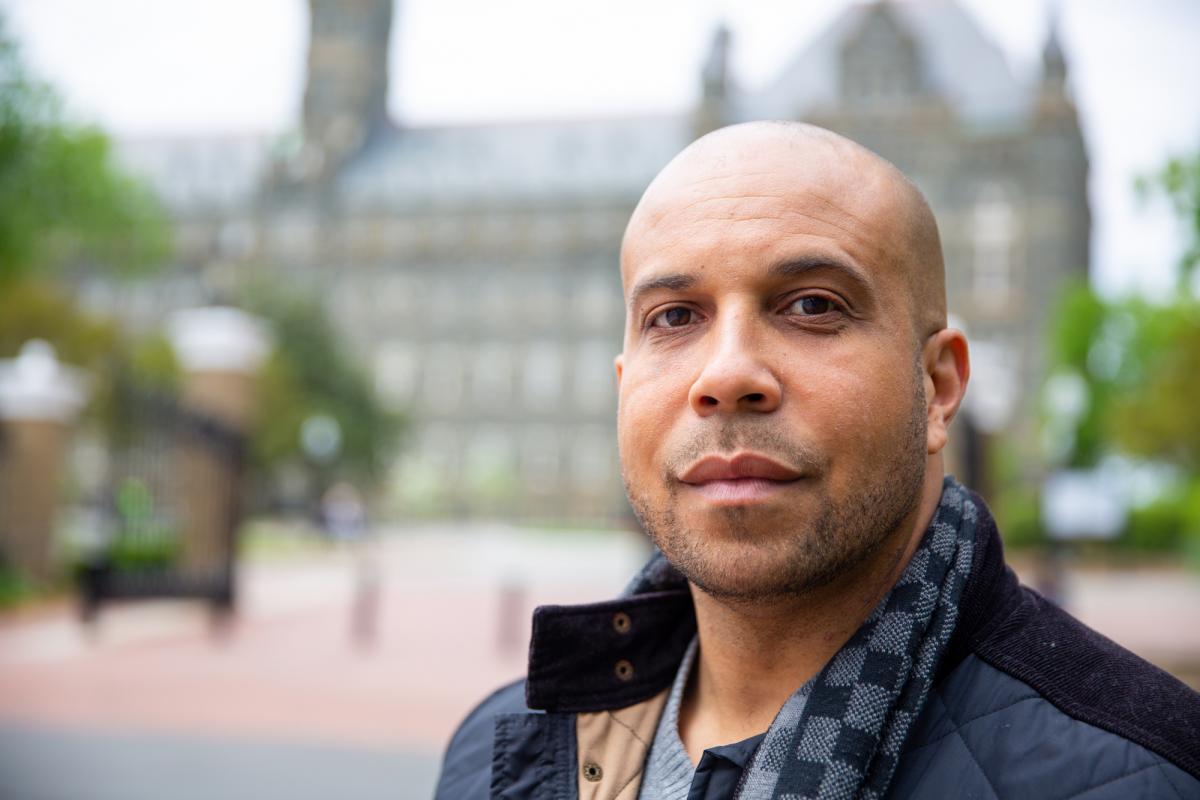 Brian Ferguson, a black man wearing a jacket stands on Georgetown's campus.