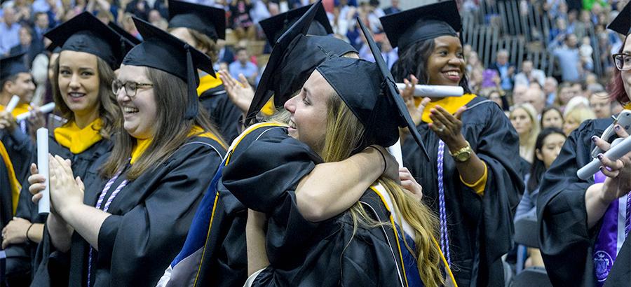 Two graduates hug among a crowd of celebrating graduates.