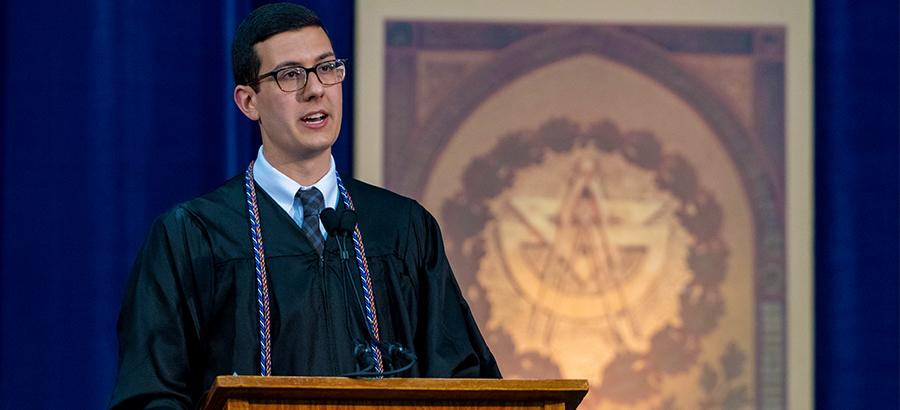 Daniel Wassim, a white man wearing academic regalia, speaks from the commencement podium.