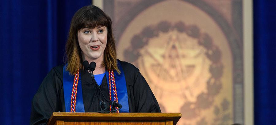 Cristine Pedersen, a white woman wearing academic regalia, speaks from the commencement podium.
