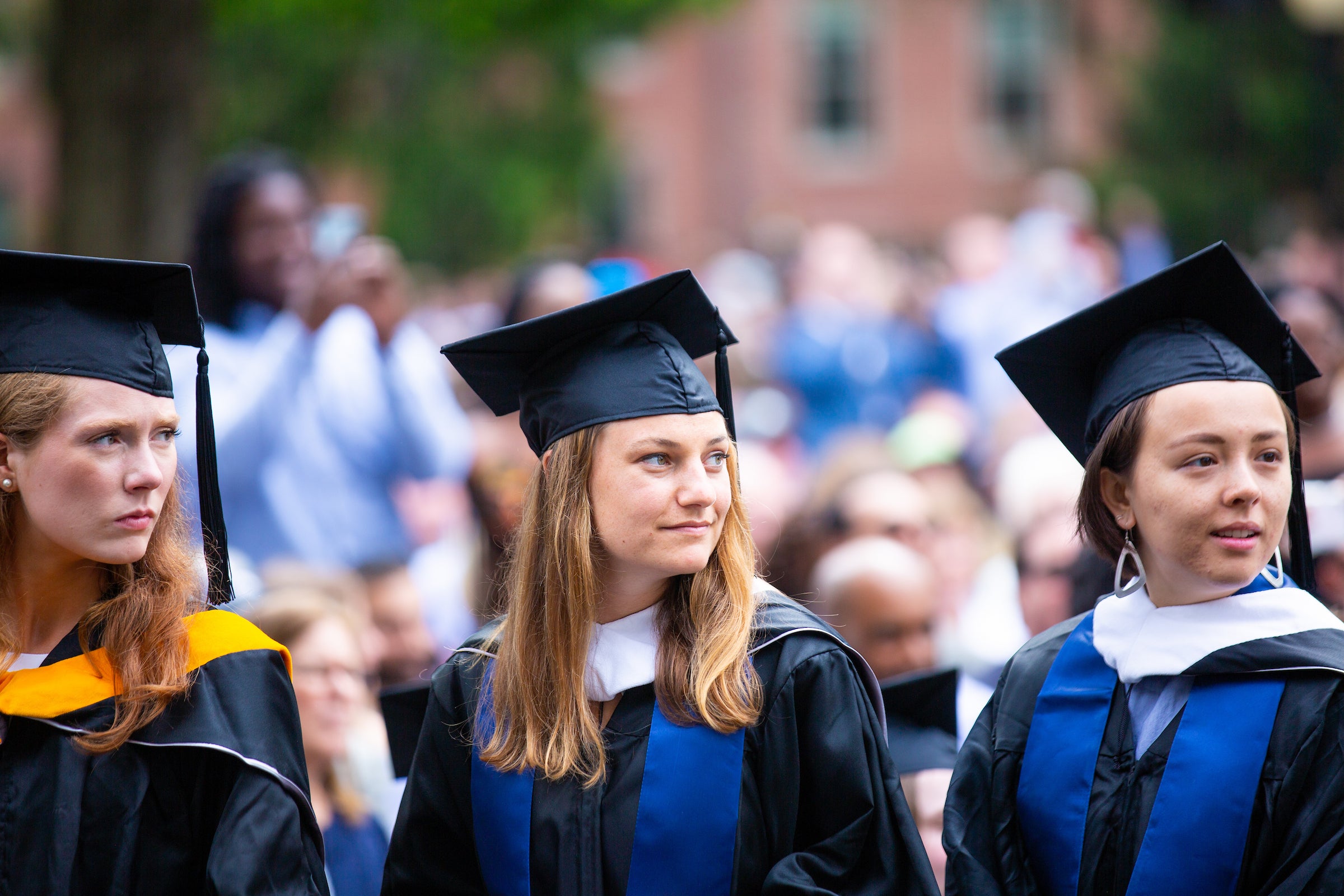 Three women, dressed in academic regalia, stand among a crowd of people.