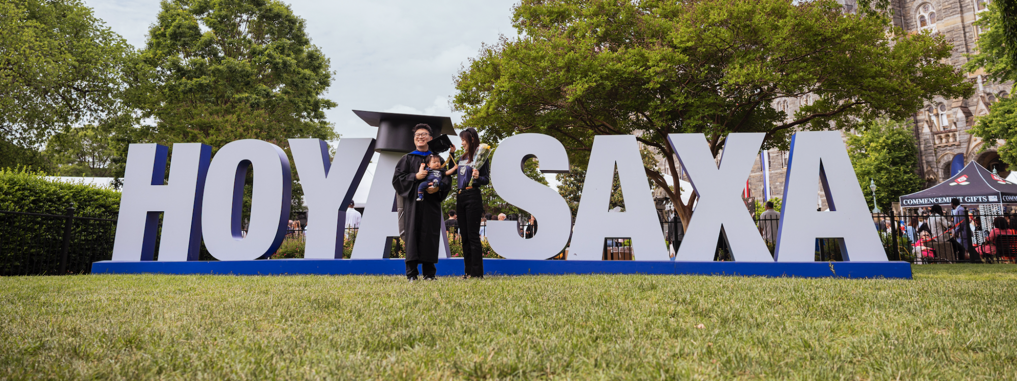 A graduate with his partner and baby stand on Healy Lawn in front of a giant sign reading 